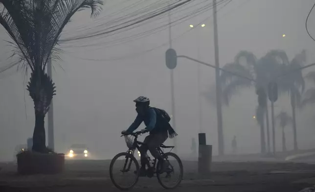 A cyclist pedals through the smoke in Brasilia, Brazil, coming from a fire in the environmentally protected area of Brasilia National Park, early Monday, Sept. 16, 2024. The head of the agency that manages protected areas, Mauro Pires, told the local press that the fire is man-made and appears to have started near the edge of the presidency's official countryside residence. (AP Photo/Eraldo Peres)