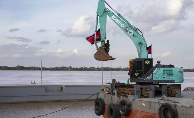 A worker raises a Cambodian flag with the help of an excavator on a barge by the mouth of a canal, at Prek Takoe village, Phnom Penh, Cambodia, Tuesday, July 30, 2024. (AP Photo/Anton L. Delgado)