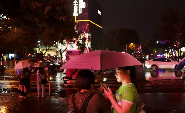 FILE - People look at their phones while waiting to cross an intersection in the rain at the Taikoo Li Sanlitun shopping center in Beijing, July, 30, 2024. (AP Photo/Vincent Thian, File)