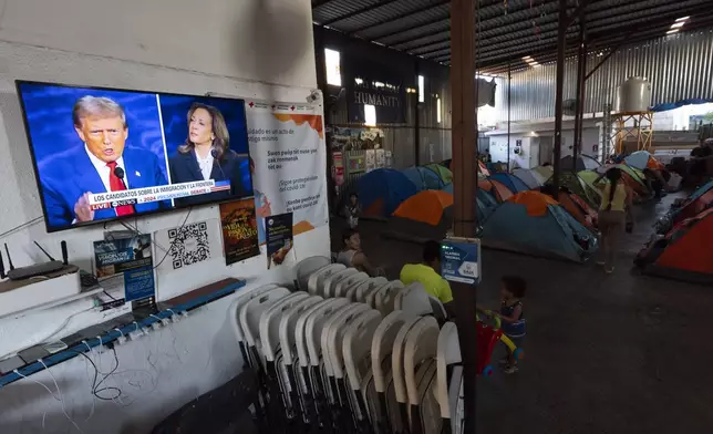 An asylum-seeking migrant from Michoacan, Mexico, walks back to the tent she shares with her family as the presidential debate between Republican presidential nominee former President Donald Trump and Democratic presidential nominee Vice President Kamala Harris plays on a television Tuesday, Sept. 10, 2024, at a shelter for migrants in Tijuana, Mexico. (AP Photo/Gregory Bull)