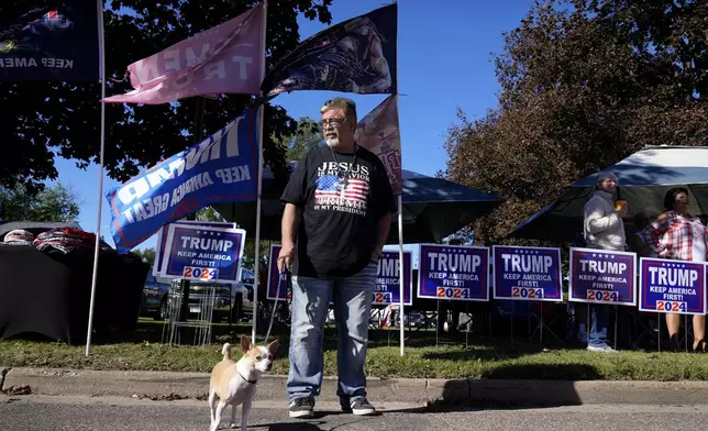 Merchandise vendor Sam Smith, of Sarasota, Fla., stands with his dog Milo while waiting for Republican presidential nominee former President Donald Trump to arrive at a rally, Saturday, Sept. 28, 2024, in Prairie du Chien, Wis. (AP Photo/Charlie Neibergall)