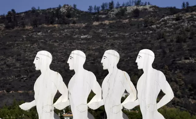 A sculpture, depicting marathon runners, stands in front of a blackened hill which had been burned in a mid-August wildfire, at Dionysos northern suburb of Athens, Thursday, Aug. 22, 2024. (AP Photo/Thanassis Stavrakis)