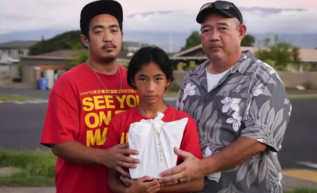 FILE - Briena Mae Rabang, 10, holds the ashes of her great-grandmother Sharlene Rabang, who was named as the 100th victim of the Lahaina wildfire, while posing for a photo with her father Branden, left, and grandfather Brandon, right, Friday, Dec. 8, 2023, in Kahului, Hawaii. (AP Photo/Lindsey Wasson, File)