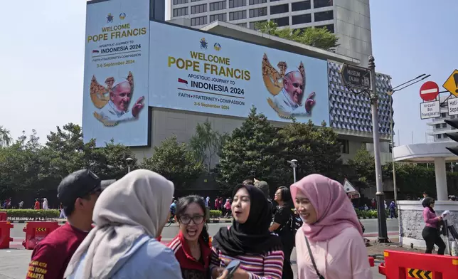 Muslim women chat as a large digital advertisement board with a welcoming message for Pope Francis is displayed on the facade of a building nearby in Jakarta, Sunday, Sept. 1, 2024, ahead of his visit to Indonesia from Sept. 3-6. (AP Photo/Dita Alangkara)