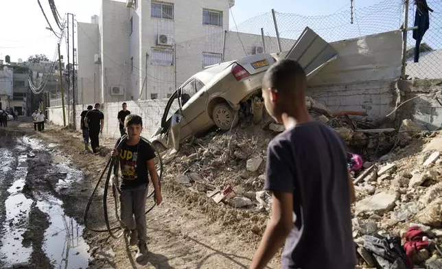 Palestinian refugees walk past a damaged vehicle in the West Bank refugee camp of Tulkarem, during an Israeli army operation in Tulkarem, Thursday, Sept. 12, 2024. (AP Photo/Nasser Nasser)