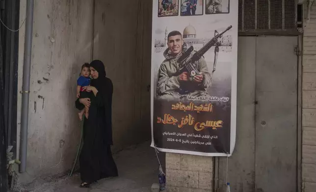 A woman carrying her child passes a banner with a picture of Issa Jallad, 17, who was killed by Israeli forces during a raid two days earlier in Jenin, West Bank, Saturday, June 8, 2024. (AP Photo/Bram Janssen)