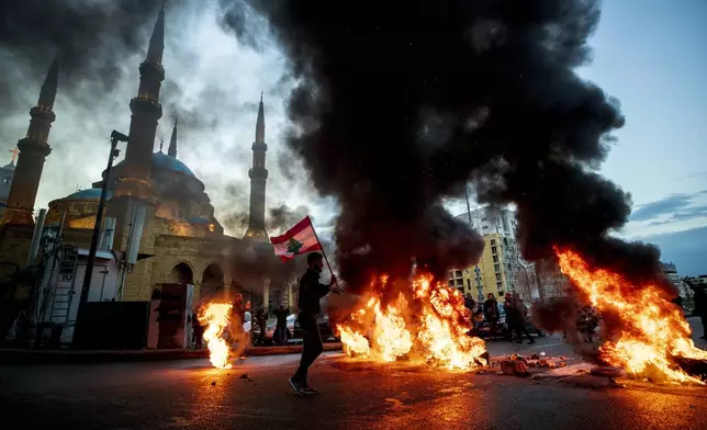 FILE - A protester holds up a Lebanese national flag as he walks in front of burning tires that are blocking a main road, during a protest in downtown Beirut, on March 3, 2021. (AP Photo/Hassan Ammar, File)