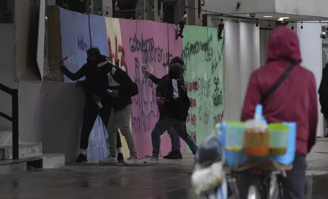 Masked youths crack a window as others spray paint anti-government slogans on a barrier protecting a storefront restaurant during a march marking the 10-year anniversary of the disappearance of 43 students from an Ayotzinapa rural teacher's college, in Mexico City, Thursday, Sept. 26, 2024. (AP Photo/Fernando Llano)