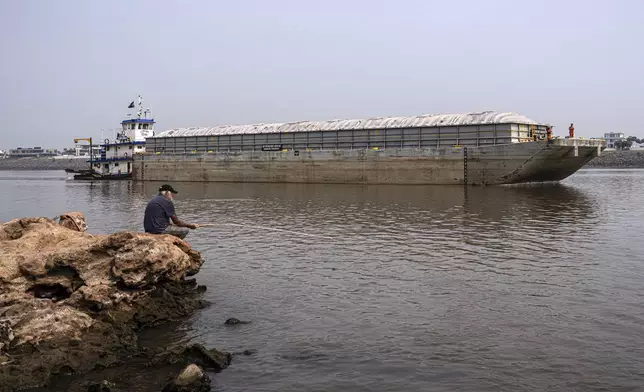 A man fishes on the shore of the Paraguay River where a tugboat pushes a barge amid low water levels and a drought in Mariano Roque Alonso, Paraguay, Monday, Sept. 9, 2024. (AP Photo/Jorge Saenz)