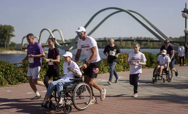 A man pushes a woman in a wheelchair during a charity run to raise awareness for people with disabilities as they pass Wave pedestrian bridge in Kyiv, Ukraine, Saturday, Sept. 14, 2024. (AP Photo/Evgeniy Maloletka)