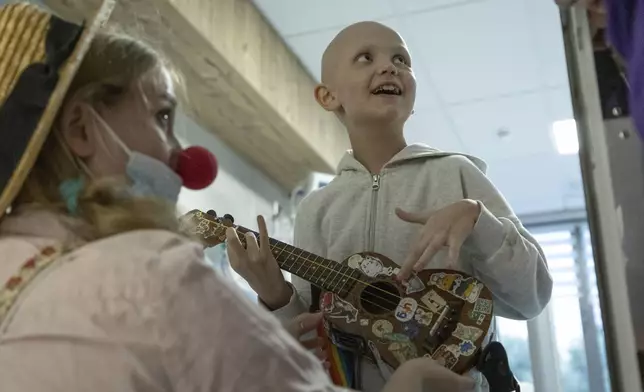 Tetiana Nosova, who goes by the clown name of Zhuzha, a volunteer from the "Bureau of Smiles and Support" watches as Kira Vertetska, 8, plays a ukulele at Okhmatdyt children's hospital in Kyiv, Ukraine, Thursday Sept. 19, 2024. (AP Photo/Anton Shtuka)