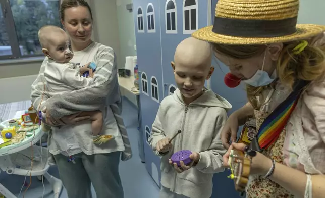 Tetiana Nosova, who goes by the clown name of Zhuzha, a volunteer from the "Bureau of Smiles and Support" plays a ukulele as she stands with Michael Bilyk, who is held by his mother Antonina Malyshko, and Kira Vertetska, 8, at Okhmatdyt children's hospital in Kyiv, Ukraine, Thursday Sept. 19, 2024. (AP Photo/Anton Shtuka)