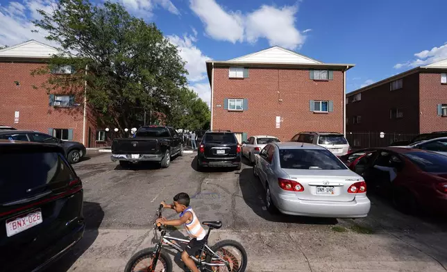 A boy guides his bicycle past apartment buildings as a rally staged by the East Colfax Community Collective is held in the courtyard to address chronic problems in the apartment buildings occupied by people displaced from their home countries in central and South America Tuesday, Sept. 3, 2024, in Aurora, Colo. (AP Photo/David Zalubowski)