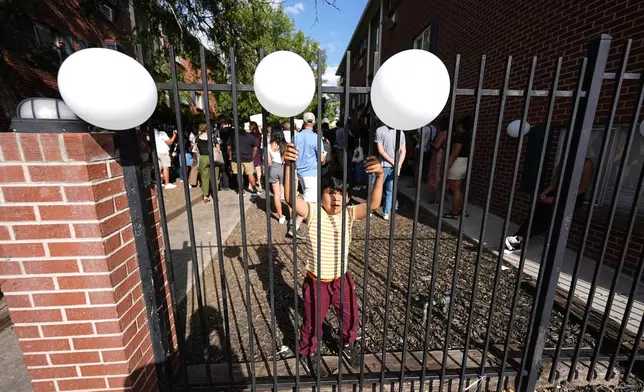 A young boy climbs an iron fence to reach balloons during a rally staged by the East Colfax Community Collective to address chronic problems in the apartment buildings occupied by people displaced from their home countries in central and South America Tuesday, Sept. 3, 2024, in Aurora, Colo. (AP Photo/David Zalubowski)