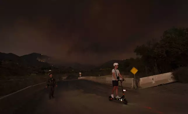 An Orange County Sheriff's officer escorts a man on a scooter out of a closed area as the Airport Fire burns, behind, Monday, Sept. 9, 2024, in Trabuco Canyon, Calif. (AP Photo/Gregory Bull)