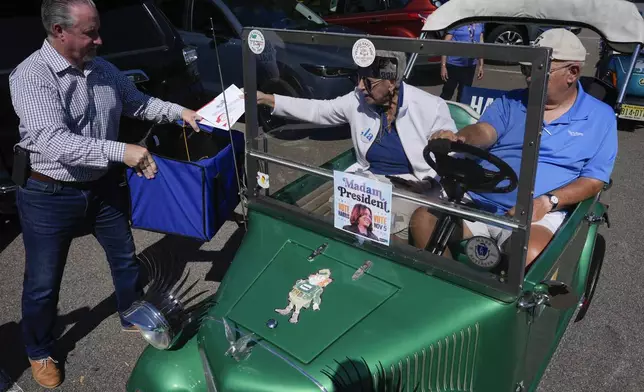 A couple drops their mail-in ballots into a collection box held by Sumter County Supervisor of Elections Bill Keen, during a golf cart parade to deliver mail-in votes organized by the Villages Democratic Club, in The Villages, Fla., Monday, Oct. 14, 2024. (AP Photo/Rebecca Blackwell)