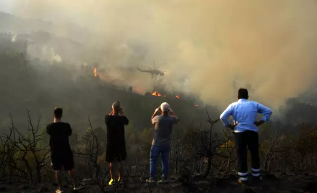 People watch and take photos of a helicopter dropping water on the flames during a third day of a wildfire, in Sofiana village, about 142 kilometers (88 miles) west of Athens, Greece, on Tuesday, Oct. 1, 2024. (AP Photo/Petros Giannakouris)