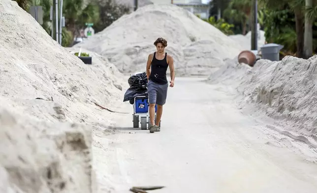 David DeMeza walks out with belongings through sands pushed on to the streets by Hurricane Helene, Wednesday, Oct. 2, 2024, in Treasure Island, Fla. (AP Photo/Mike Carlson)