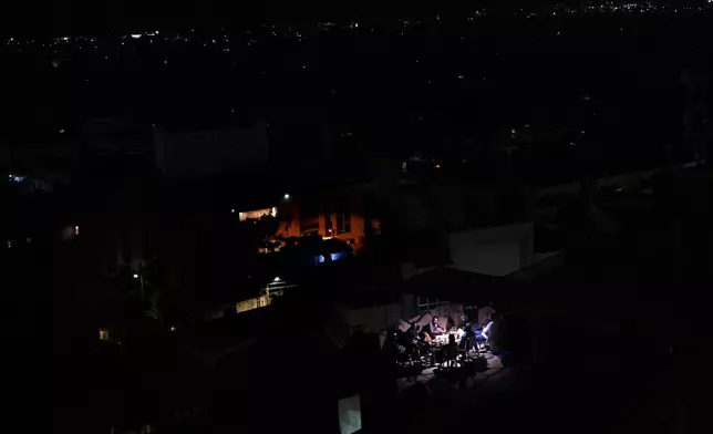 Residents sit on the roof of a building and have dinner as Dahiyeh suburb, background, remains in darkness after Israeli airstrikes, Lebanon, Friday, Oct. 11, 2024. (AP Photo/Hassan Ammar)