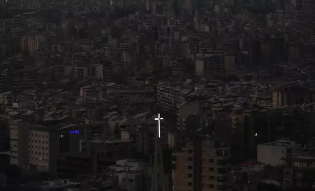 The cross atop of Our Lady of Hadath Church appears in front of Beirut's Dahiyeh suburb, background, that remains in darkness after Israeli airstrikes, Lebanon, Wednesday, Oct. 16, 2024. (AP Photo/Hassan Ammar)