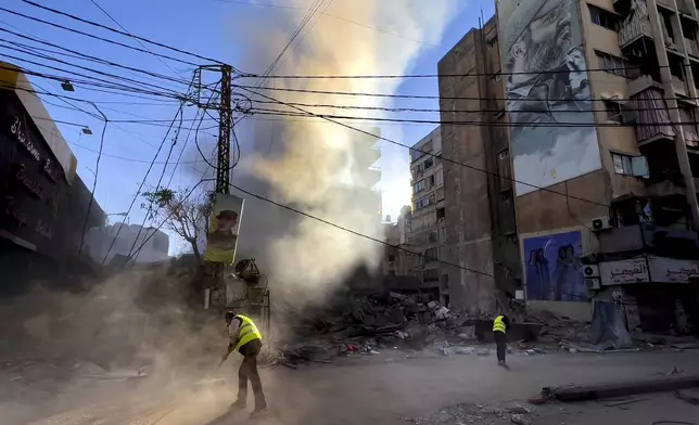 Workers clean a street under a giant portrait of the late Hezbollah military commander Imad Mughniyeh, as smoke rises from a destroyed building that was hit by an Israeli airstrike in Dahiyeh, in the southern suburb of Beirut, Lebanon, Sunday, Oct. 20, 2024. (AP Photo/Hussein Malla)