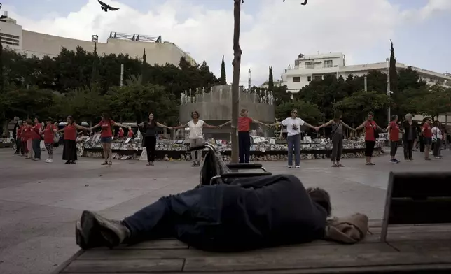 A man sleeps on a bench as people hold hands at a memorial marking the one-year anniversary of the Hamas attack on Israel, in Tel Aviv, Monday, Oct. 7, 2024. (AP Photo/Oded Balilty)