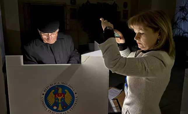 An election official assists a voter with her mobile phone flashlight during a power failure in Chisinau, Moldova, Sunday, Oct. 20, 2024, during a presidential election and a referendum on whether to enshrine in the Constitution the country's path to European Union membership. (AP Photo/Nicolae Dumitrache)