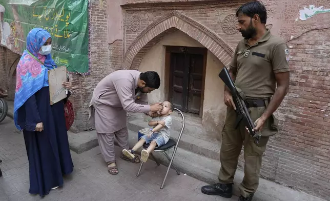 A police officer stands guard as a health worker, center, administers a polio vaccine to a child at a neighbourhood of Lahore, Pakistan, Monday, Oct. 28, 2024. (AP Photo/K.M. Chaudary)