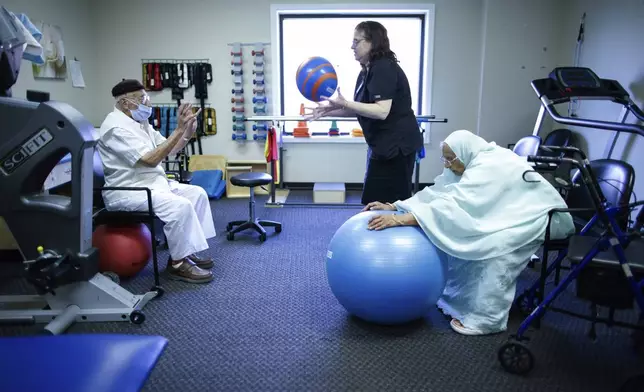 Mohamed, 84, left, receives physical therapy with therapist Susan Fenyes, center, while his wife, Bibi, 84, stretches with a ball at Sunshine Adult Day Center in Bergenfield, N.J., Monday, Aug. 26, 2024. (AP Photo/Kena Betancur)