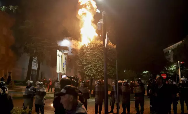 Fire burns behind a riot police cordon during an anti-government rally set up by the opposition, in Tirana, Albania, Monday, Oct. 7, 2024. (AP Photo/Hameraldi Agolli)