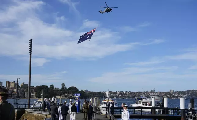 A helicopter flies with a giant Australian flag over the harbor during Britain's King Charles III and Queen Camilla visit at the Sydney Opera House in Sydney, Australia, Tuesday, Oct. 22, 2024. (AP Photo/Mark Baker, Pool)