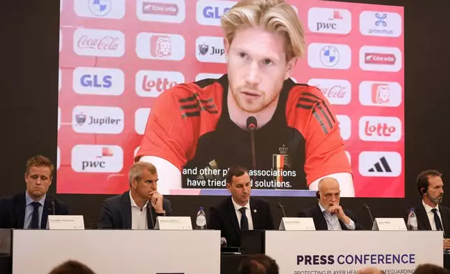 Soccer player Kevin De Bruyne is shown on a large screen, as panel members, from left to right, NISO executive president Kristoffer Vatshaug, EPL director of international football relations, EU affairs Mathieu Moreuil, FIFPRO Europe president David Terrier, LaLiga president Javier Tebas and AIC resident Umberto Calcagno listen during a media conference on protecting player health and safeguarding the sustainability of national leagues at the Residence Palace in Brussels, on Monday, Oct. 14, Oct. 8, 2024. (AP Photo/Virginia Mayo)