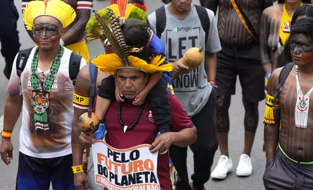 An Indigenous father carries his son on his shoulder, holding a sign with a message that reads in Portuguese: "For the Planet's Future", during a protest against a proposed constitutional amendment that threatens some Indigenous land rights, in Brasilia, Brazil, Wednesday, Oct. 30, 2024. (AP Photo/Eraldo Peres)