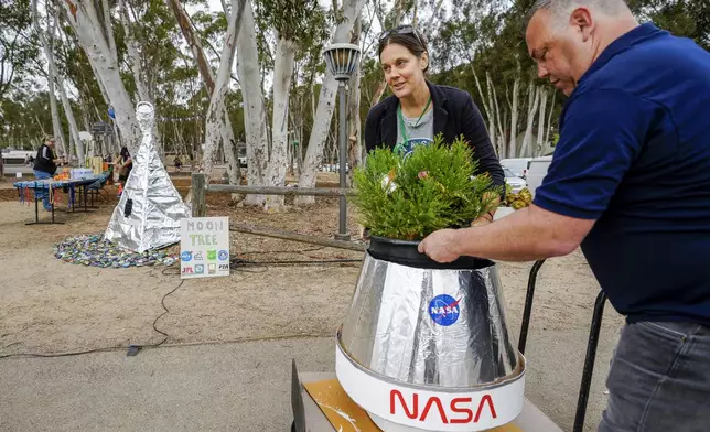 Santiago parent volunteer Stacie Aguesse, left, delivers a small Giant Sequoia tree from NASA's Artemis I Mission's tree seeds that traveled around the moon twice, as NASA scientists, JPL engineers, US Forest Service representatives, and teachers join Santiago STEAM Magnet Elementary School students at a ceremony to plant it after the school was honored in the spring of 2024 to become NASA Moon Tree Stewards in Lake Forest, Calif., on Monday, Oct. 14, 2024. (AP Photo/Damian Dovarganes)