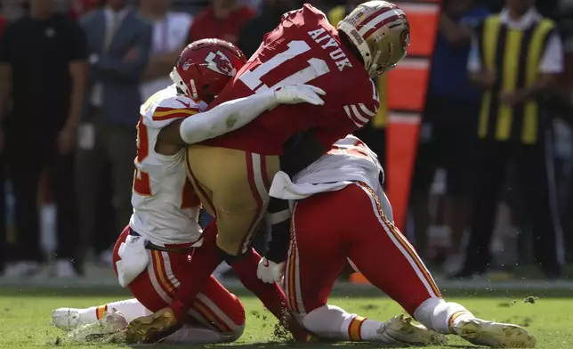 San Francisco 49ers wide receiver Brandon Aiyuk (11) is hit by Kansas City Chiefs cornerback Trent McDuffie, left, and safety Chamarri Conner during the first half of an NFL football game in Santa Clara, Calif., Sunday, Oct. 20, 2024. (AP Photo/Jed Jacobsohn)