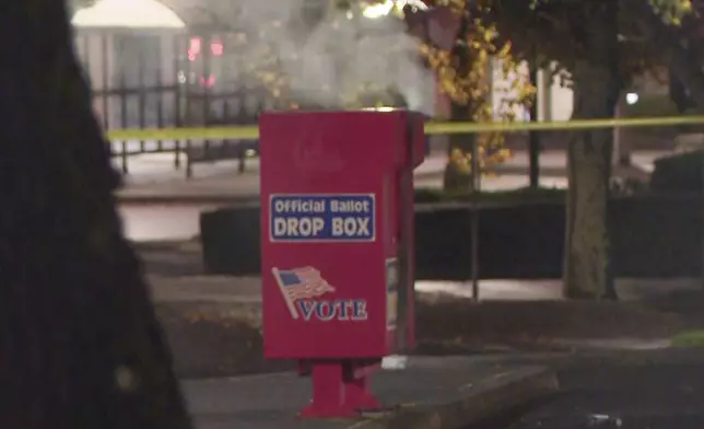 In this image made from a video provided by KGW8, authorities investigate the scene as smoke pours out of a ballot box on Monday, Oct. 28, 2024, in Vancouver, Wash. (KGW8 via AP)