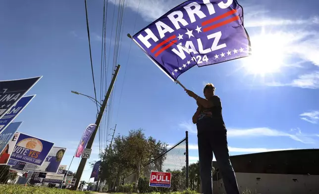 Metrowest resident Cynthia Ramirez waves a Harris-Walz campaign flag while waving to voters in line during early voting at the Orange County Supervisor of Elections precinct on Kaley Avenue in Orlando, Fla., Friday, Oct. 25, 2024. (Joe Burbank/Orlando Sentinel via AP)