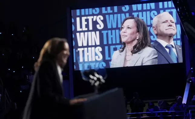 Democratic presidential nominee Vice President Kamala Harris, center, and her running mate Minnesota Gov. Tim Walz, right, appear on screen as Harris speaks at a campaign rally at East Carolina University in Greenville, N.C., Sunday, Oct. 13, 2024. (AP Photo/Susan Walsh)