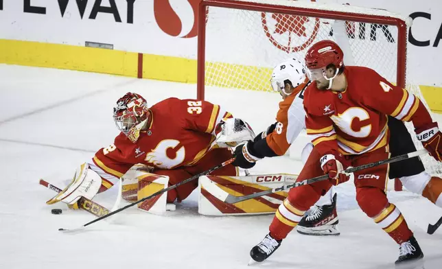 Philadelphia Flyers' Morgan Frost, center, stretches for the puck as Calgary Flames' Rasmus Andersson, right, checks him into goalie Dustin Wolf during the first period of an NHL hockey game in Calgary Alberta, Saturday, Oct. 12, 2024. (Jeff McIntosh/The Canadian Press via AP)