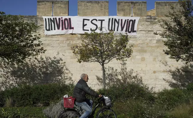 FILE - A man rides a bicycle in front of a banner reading: "A rape is a rape," in Avignon, southern France, on Oct. 16, 2024. (AP Photo/Lewis Joly, File)