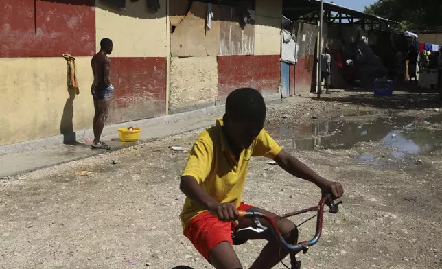 A youth cycles past a man taking a bucket bath at a school where people displaced by gang violence have been living for over a year in Port-au-Prince, Haiti, Friday, Sept. 20, 2024. (AP Photo/Odelyn Joseph)