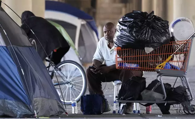 A man in a homeless encampment sits among possessions after Louisiana State police gave instructions for them to move to a different pre-designated location as they perform a sweep in advance of a Taylor Swift concert in New Orleans, Wednesday, Oct. 23, 2024. (AP Photo/Gerald Herbert)