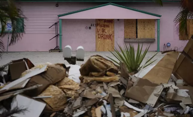 A boarded up business, marked with graffiti reading "Go home Milton, U R drunk," is seen past debris from Hurricane Helene flooding piled up outside a home, ahead of the arrival of Hurricane Milton, in Holmes Beach on Anna Maria Island, Fla., Tuesday, Oct. 8, 2024. (AP Photo/Rebecca Blackwell)