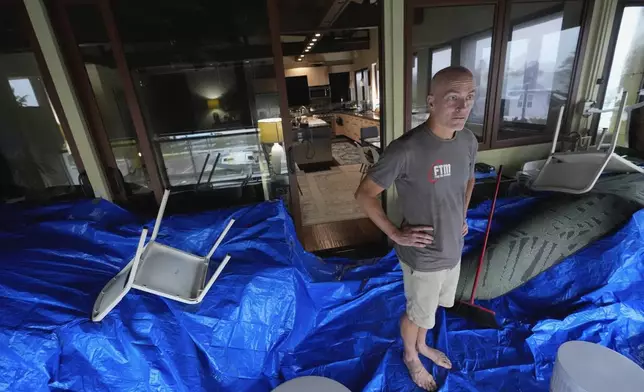 Christian Burke stands on the third floor of his home, where he, his mother, and his aunt plan to ride out Hurricane Milton with views through hurricane proof glass over Tampa Bay, in Gulfport, Fla., Wednesday, Oct. 9, 2024. Burke, who said his engineer father built the concrete home to withstand a Category 5 hurricane, expects his home to turn into an island, with up to 8 feet of water filling the raised first floor. Behind Burke, a window reflects a boat deposited by Hurricane Helene in the bay front park across the street. (AP Photo/Rebecca Blackwell)