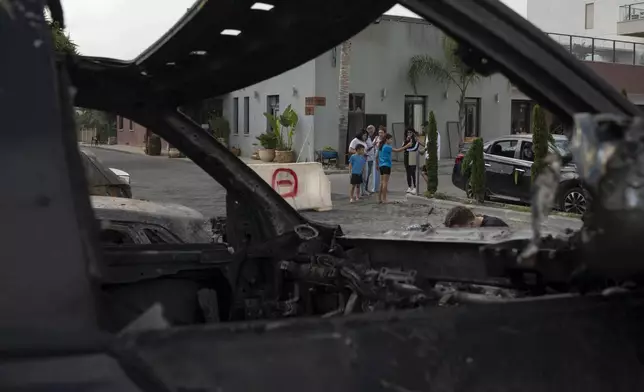 People stand next to burnt and damaged cars after a rocket, launched from Lebanon, hit an area in Kfar Vradim, northern Israel, Monday, Oct. 7, 2024. (AP Photo/Leo Correa)