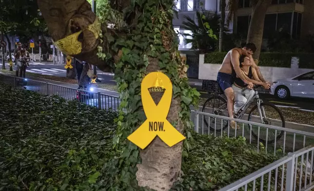 A couple rides a bicycle near a yellow ribbon sign calling for the release of hostages held in the Gaza Strip by the Hamas militant group for nearly a year, in Tel Aviv, Israel, Monday, Sept. 10, 2024. (AP Photo/Ohad Zwigenberg)