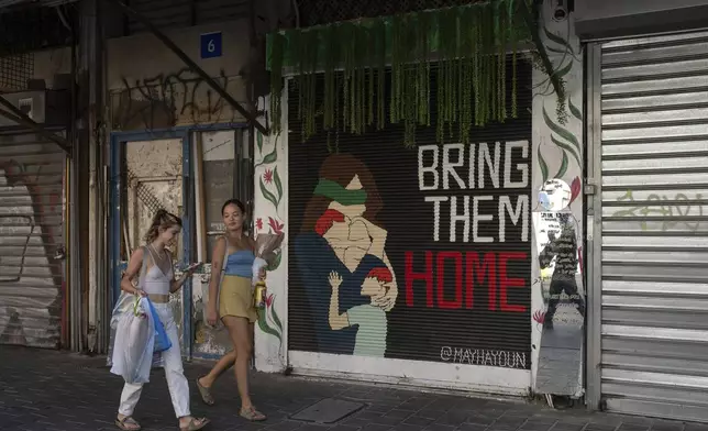 Women walk past graffiti calling for the release of hostages held by Hamas militants in the Gaza Strip for nearly a year, in the Carmel market Tel Aviv, Israel, Friday, Sept. 27, 2024. (AP Photo/Ohad Zwigenberg)