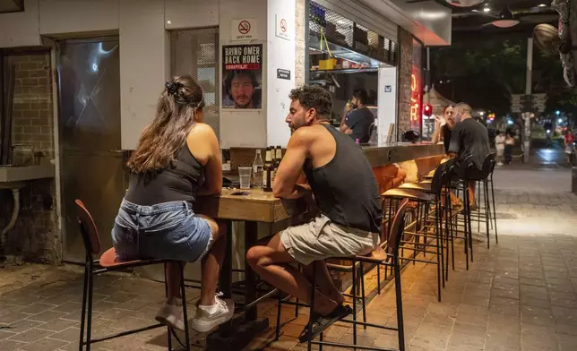 People sit in a bar near a sign calling for the release of hostages held in the Gaza Strip by the Hamas militant group for nearly a year, in Tel Aviv, Israel, Monday, Sept. 10, 2024. (AP Photo/Ohad Zwigenberg)