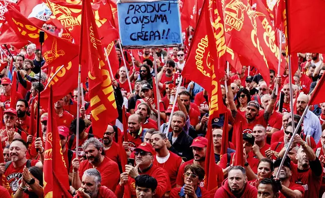 Demonstrators wave union flags on the occasion of a national strike of metalworkers of the automotive sector for the first time in 20 years, in Rome, Friday, Oct. 18 2024. Writing on sign at center reads in Italian "Produce, Consume and Die!". (Roberto Monaldo/LaPresse via AP)