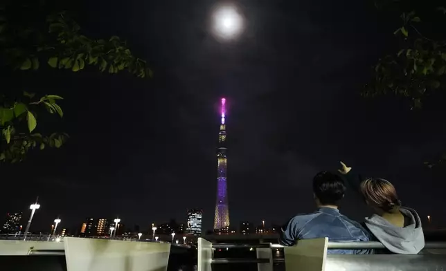 Rui Nakayama, left, and Hana Shimizu, 20-year-old college students, sit along the Sumida River as the moon shines behind Tokyo Skytree in Tokyo, Japan, Thursday, Oct. 17, 2024. (AP Photo/Hiro Komae)